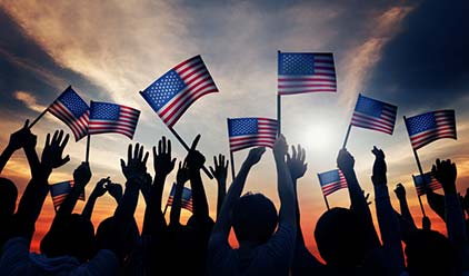 A crowd of people wave American flags against a dramatic sky.